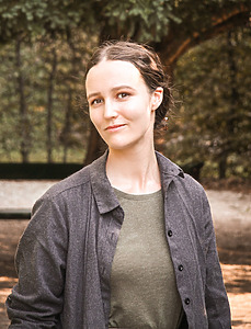 A women standing in nature background, she has dark brown hair in a bun, wearing a gray shirt and a T-shirt underneath