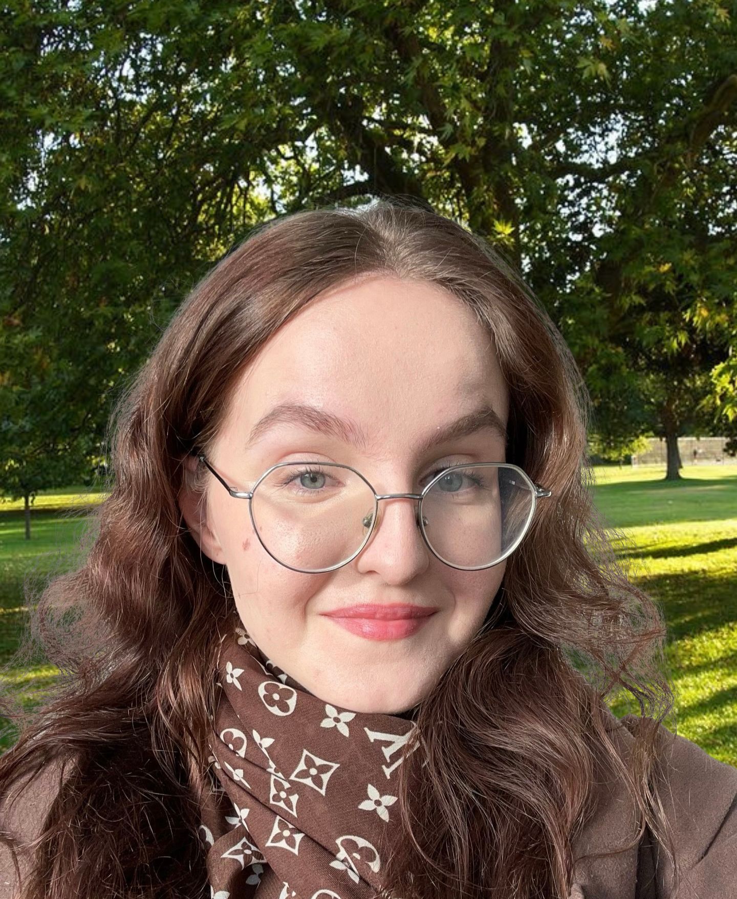 A young woman with long wavy brown hair, wearing a brown coat and a Louis Vuitton patterned scarf, stands in a park with green grass and trees in the background. She is wearing round glasses and smiling.