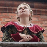 With a light, braided hair, a woman leaning into the enclosure looks into the distance, adorned with a variegated medieval dress.