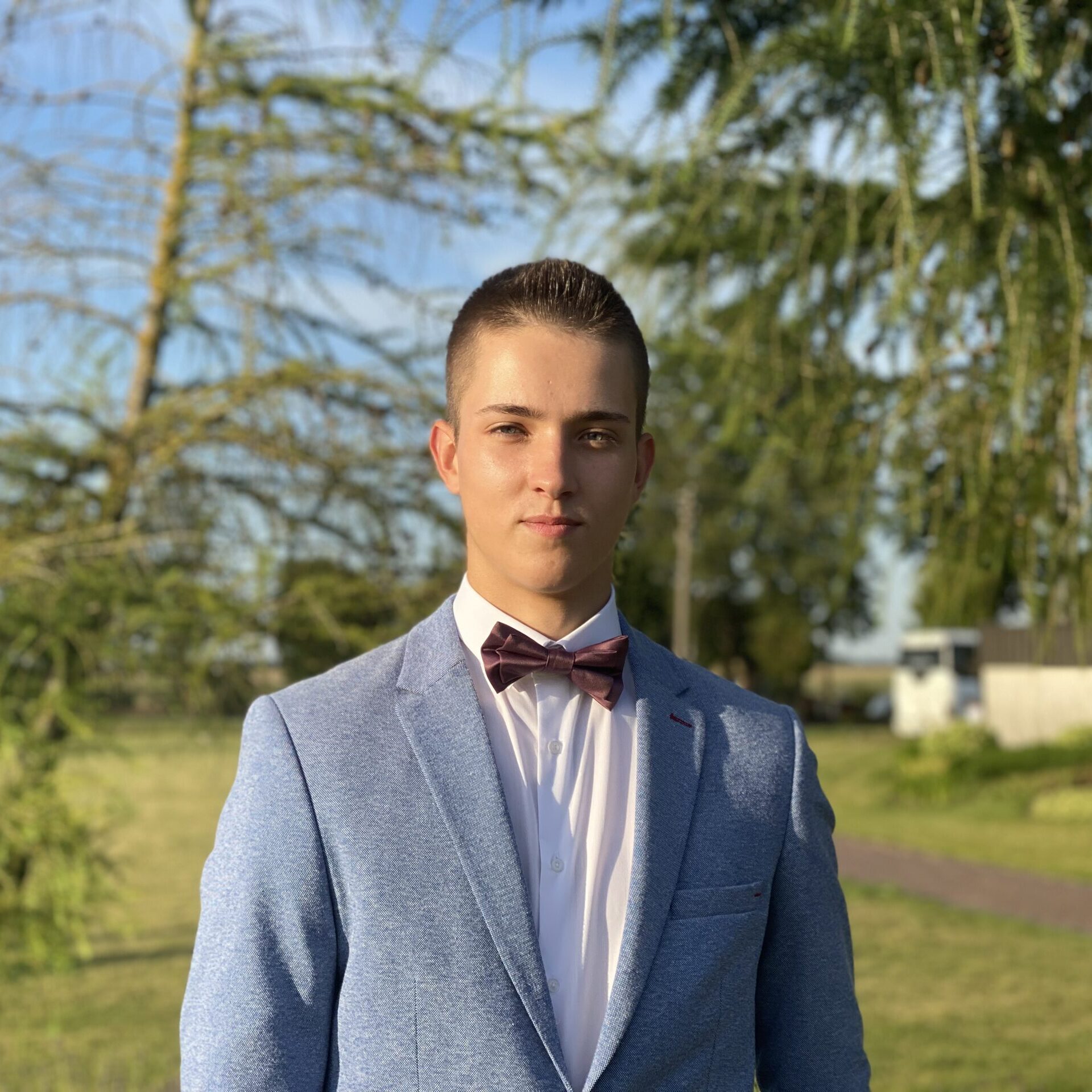 A student with short black hair wearing a shirt, a gray jacket and a frog in a forest background.