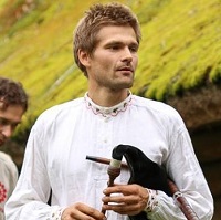 A man with blonde, short hair, in a white linen shirt, holding several musical instruments in his hands.