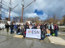 A large group of people standing by the ship and holding a white flag with a crest.