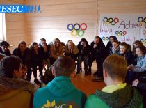 Students sitting in a circle listen to the lecturer in the middle. In the background the wall is decorated with Olympic rings