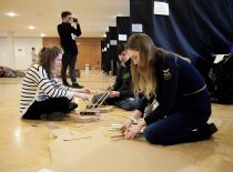 In the assembly hall, three students construct something from cardboard and wooden sticks on the ground, behind them a student is photographing something, and a student is standing far to the door.