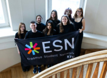 Group of people in the middle of stairs holding a flag and smilling. Behind them two windows.