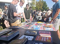 A scene from an organization fair with participants browsing a table of books and publications outdoors on a sunny day.