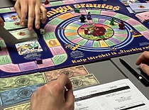 A moment from the board game "Cash Flow," showing players' hands, the game board, money, and notes.A moment from the board game "Cash Flow," showing players' hands, the game board, money, and notes.