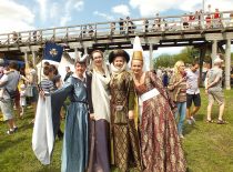 Four women dressed in high-class medieval costumes for the celebration. Behind the back of the women you can see more participants of the festival, a bridge, tents of merchants.