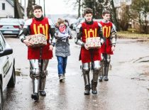 There are three students in the street dressed in armor and with a garment depicting the pillars of Gedminaičiai, two of the three students are carrying presents, and next to them is a student looking at the sheets.