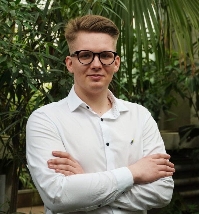 A guy standing in the middle of green backgroudn with plansts. His arms are crossed, he is wearing white shirt and glasses and he has light brown hair.