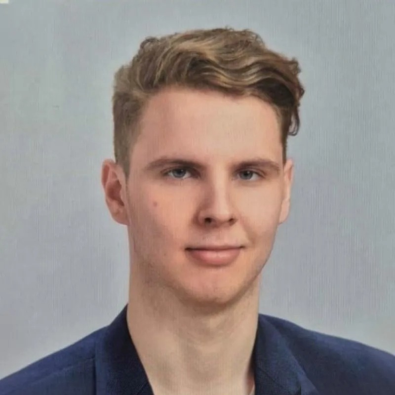 A portrait photo of a young man with short blonde hair styled with a side part. He is wearing a dark blazer and posing against a light background with a slight smile.