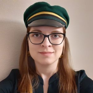 A student with long, brown hair, wearing a green hat with a yellow ribbon, glasses and a dark shirt.
