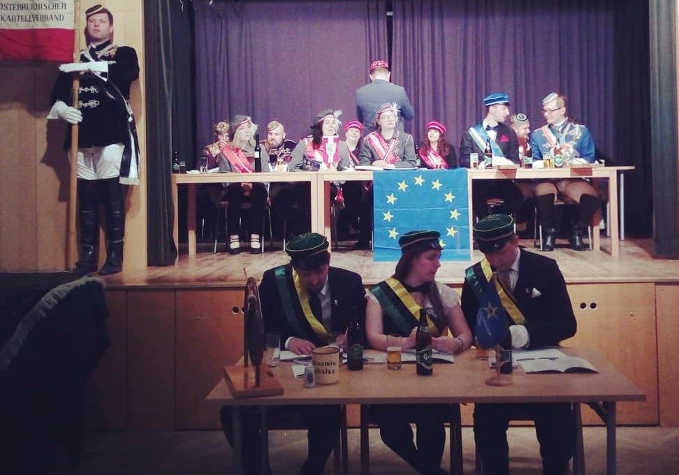 A group of students is sitting at a long table in the hall, with the European Union flag in front of the table, a student holding a poster on the left, three students sitting at the table and talking to each other.