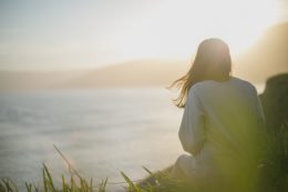 At sunset, a girl sitting by the sea, wearing a warm sweater. In the distance are mountains.
