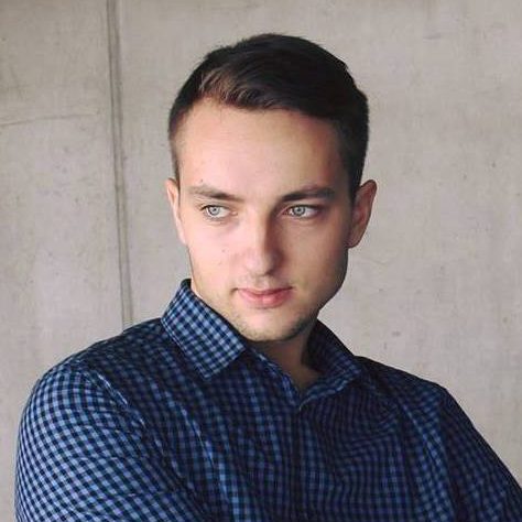 A student with short, dark hair, a dark blue checkered shirt against a concrete wall.
