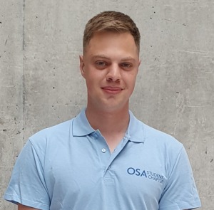 A student with short, blonde hair, in a light blue shirt on a gray background.