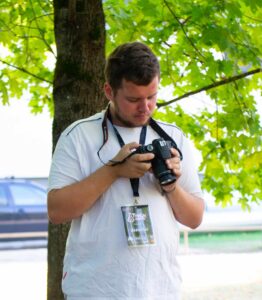A guy with white shirt standing in the middle and looking at camera. There is a green tree in the background