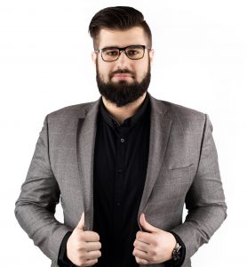 A KTU employee wearing a black shirt and gray jacket. A man with a beard, dark hair, black glasses. With both hands holds jacket's revers.