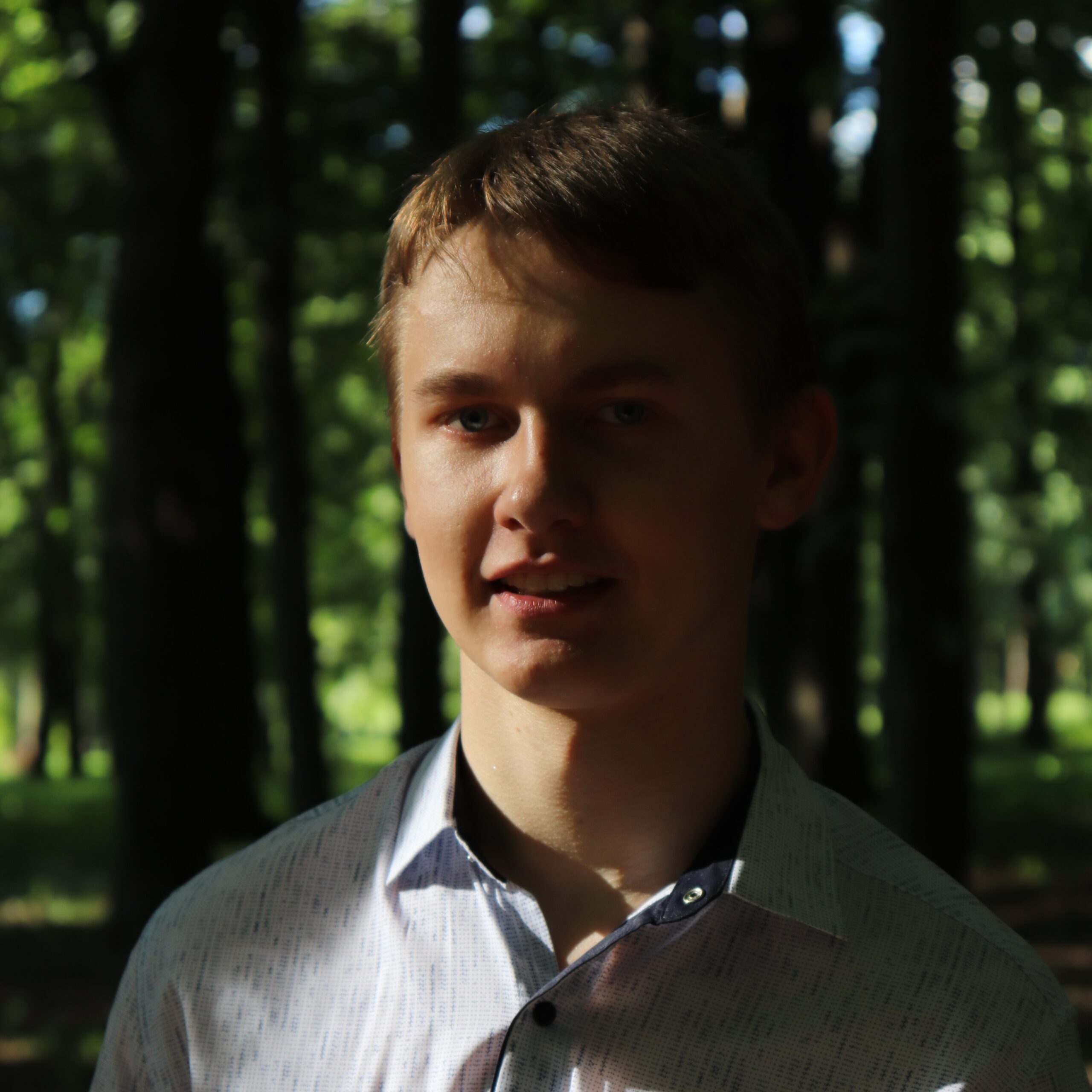 A brown hair guy wearing white shirt is standing in the middle of forest green background.