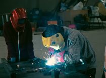 A guy with a yellow mask and a checkered shirt is bent over soldering the device, sparks can be seen. A person with a red mask is on the left side.