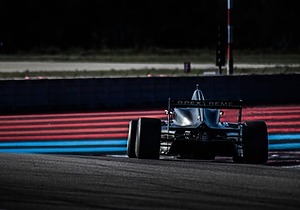 A black Formula racing car on a track with red and blue stripes, photographed from the rear. The car appears to be in motion, and the background is dark, creating a dramatic contrast.