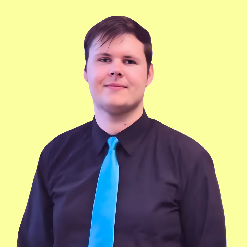 JA young man with short dark hair, wearing a black shirt and a bright blue tie, stands in front of a light yellow background and looks at the camera with a gentle smile.