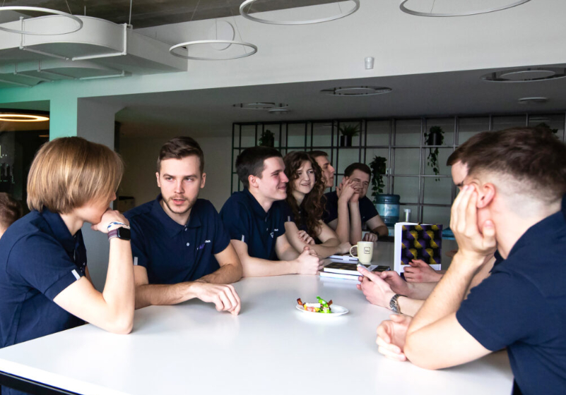Eight young adults in matching navy-blue shirts are gathered around a white table in a modern office space. They appear relaxed and engaged in conversation, with one person using a phone and others smiling; notebooks, a coffee mug, and a small plate of candy are on the table.