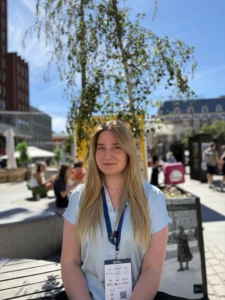 A girl with light hair, standing next to a tree in the city center.