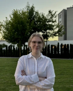 Young girl with white shirt, around her there is grass, tree and a gray building.