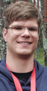 A young guy with glasses, dressed in a gray sweater and a red pendant around his neck.