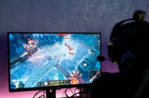 A game on the screen, a pink light at the bottom of the monitor, and a person wearing headphones next to the monitor.