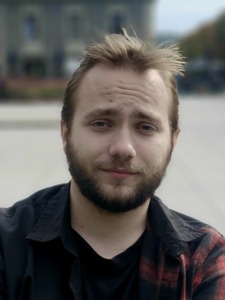 Young man with a beard. Wearing a black shirt and red with black tiles jacket.