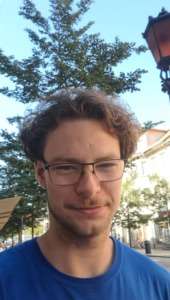 A young man in a blue T-shirt and sunglasses, behind him is a tree and a light-coloured building